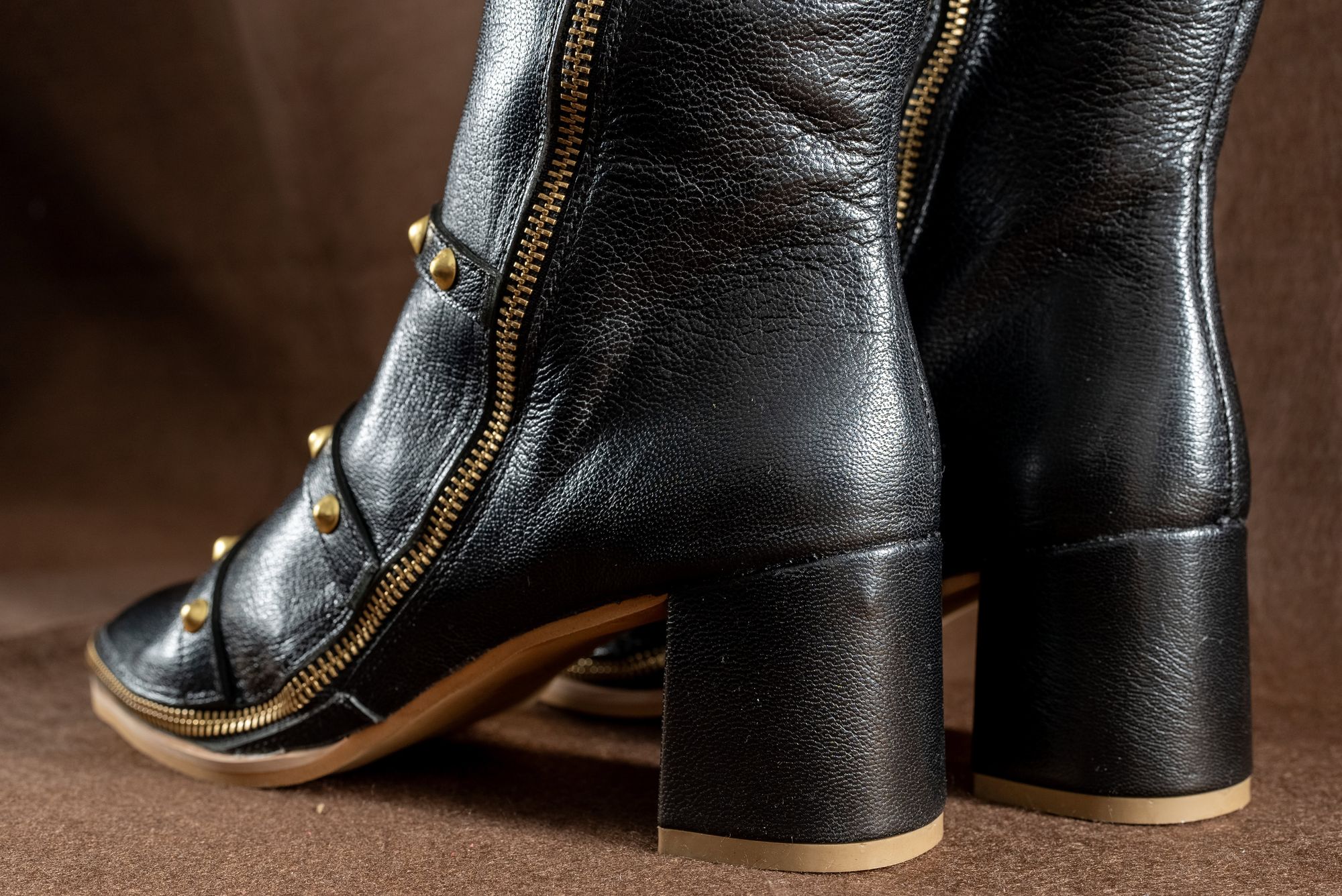 Close on black full-grain leather, set studs and a metal zip, on a warm brown ground